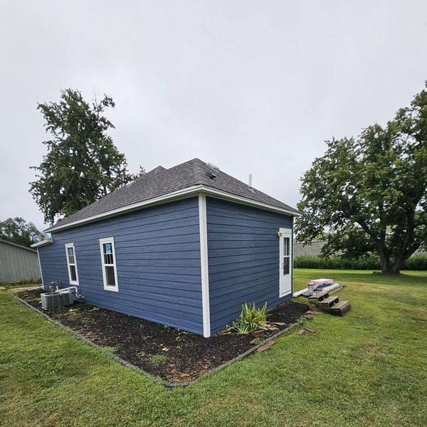 Blue vinyl-sided house with white trim and a landscaped flower bed in the yard.