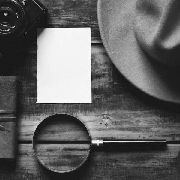 A fedora hat, magnifying glass and notepad arranged on a dark wooden table.