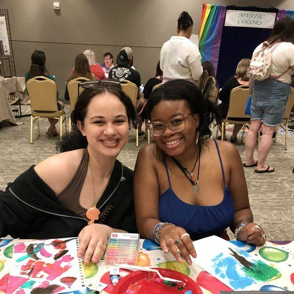 Two smiling women seated at a table covered in colorful paint and art supplies.
