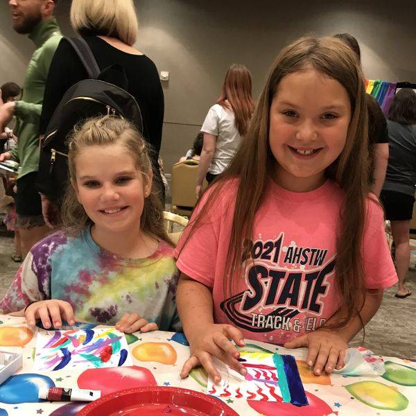 Two smiling girls in tie-dye and pink shirts at a painted art table.