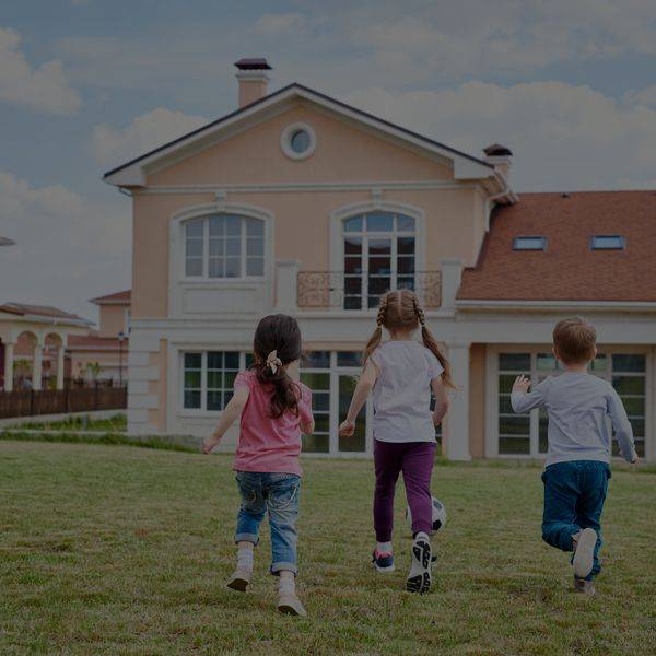 Children running across a green lawn toward a large suburban home.