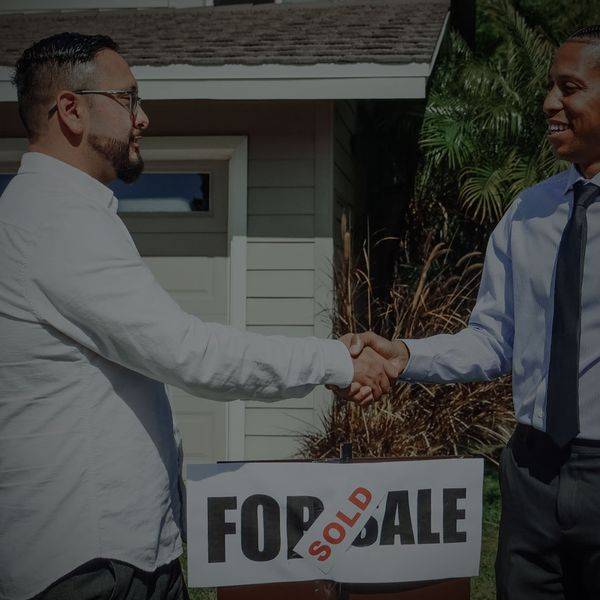 Two men shaking hands in front of a house with a For Sale sign.
