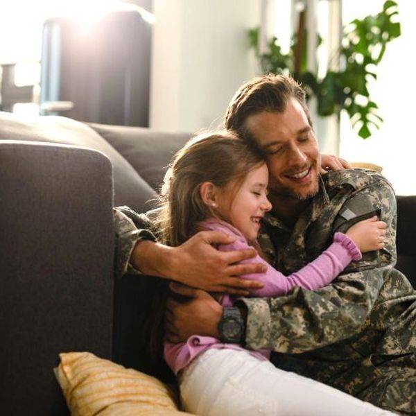 A smiling soldier in uniform hugging a young girl on a living room couch.