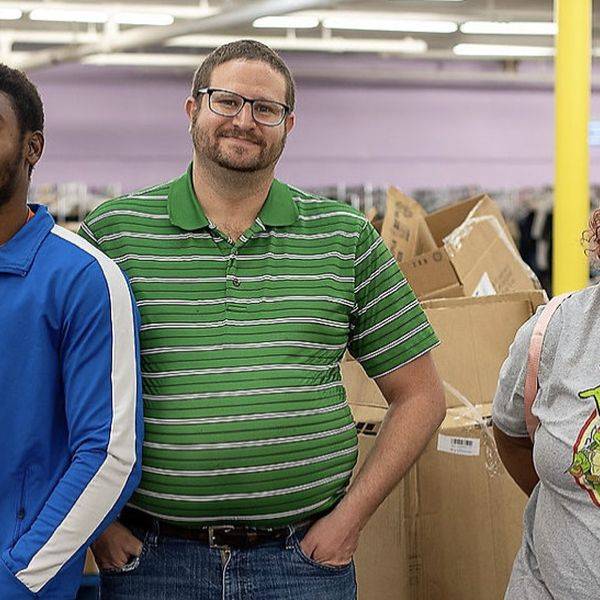 Three smiling people standing together indoors near cardboard boxes and shelves.