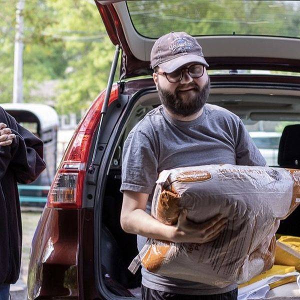 A man in a cap carrying a bag out of an open car trunk, with another person beside him.