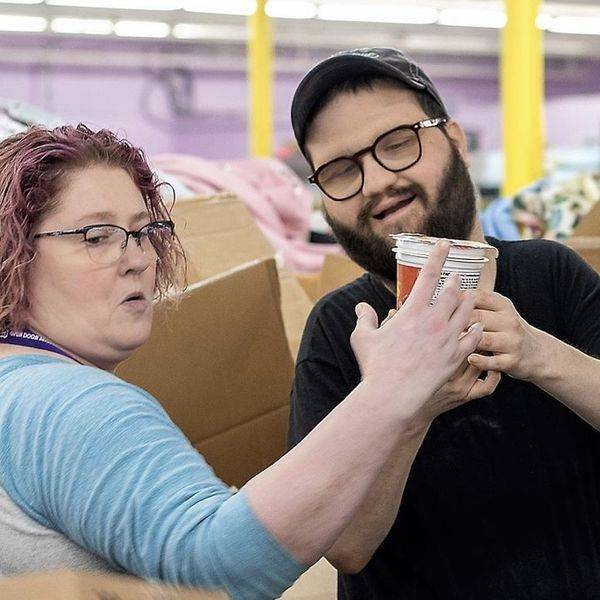 Two people smiling and holding cups in a room stacked with cardboard boxes.