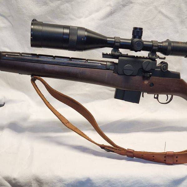 Bolt-action rifle with a scope, bipod and leather sling on white cloth.