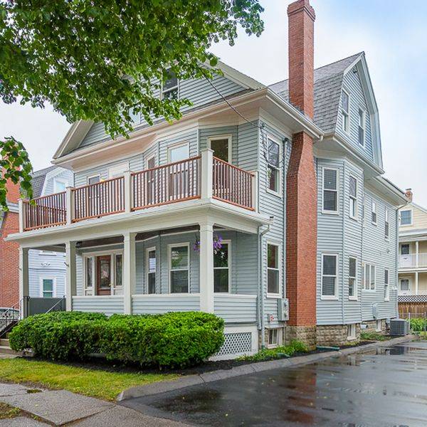 Gray two-story home with an upper balcony, brick chimney and a green hedge out front.