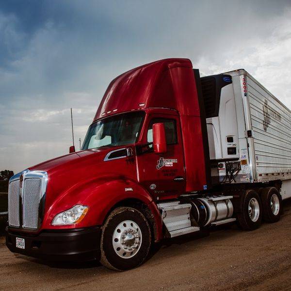 A red semi truck with a long trailer parked on a dirt lot in front of a building.