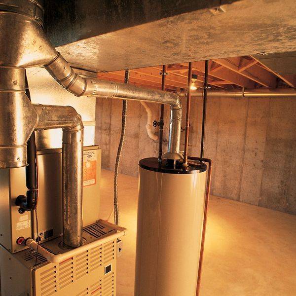 A water heater and furnace in an unfinished basement with copper pipes overhead.
