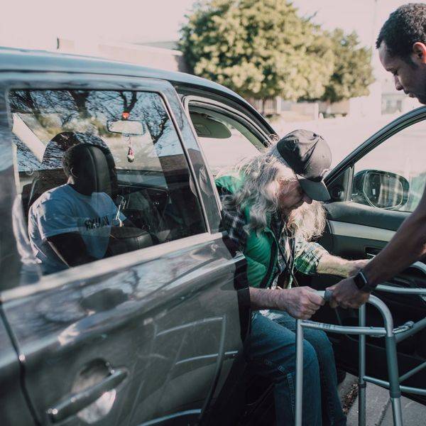 A driver helping an older passenger board a van, with a walker set up beside the door.