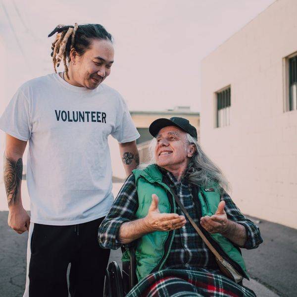 A volunteer in a white shirt walking alongside an older man on a sidewalk.