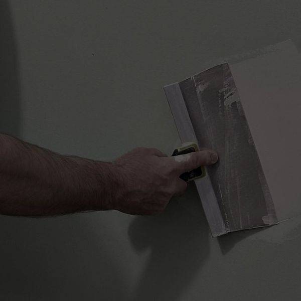 A close-up of a hand holding a taping knife against a grey wall.