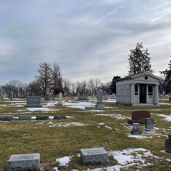 Headstones and a small white mausoleum on a snowy lawn among bare trees.