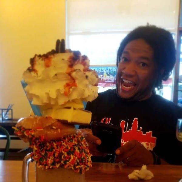 A man beside a towering loaded dessert drink with thick toppings on a restaurant table.