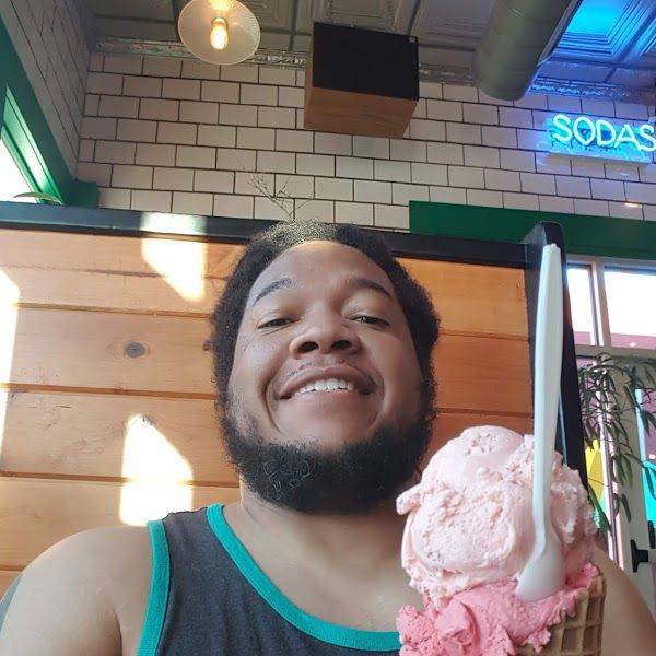 A smiling bearded man holding a waffle cone of pink and white ice cream.