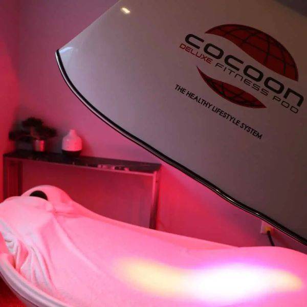 A client resting in a red-lit Cocoon Fitness Pod wellness unit with its branded side visible.