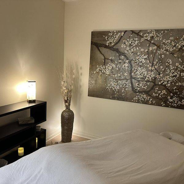 A treatment room with a made-up massage table, a lamp, and a dark framed tree-branch artwork.