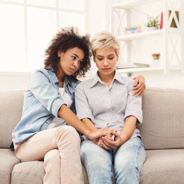 Two young women sit on a couch, one comforting the other with an arm around her.