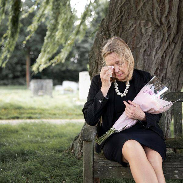 An older woman sits alone on a park bench, head in hand, holding flowers.