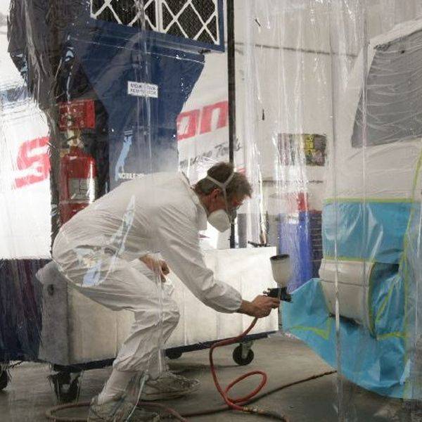 Worker in a white protective suit spraying a panel inside a mobile booth with clear plastic curtains.