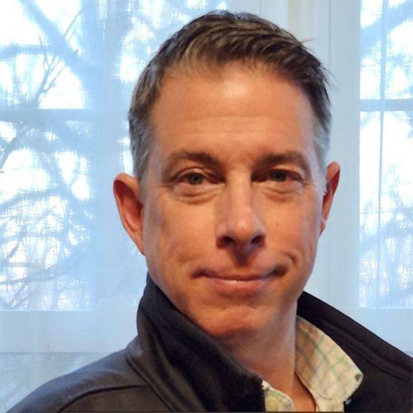 Smiling man with short hair standing near a window with bare trees outside.