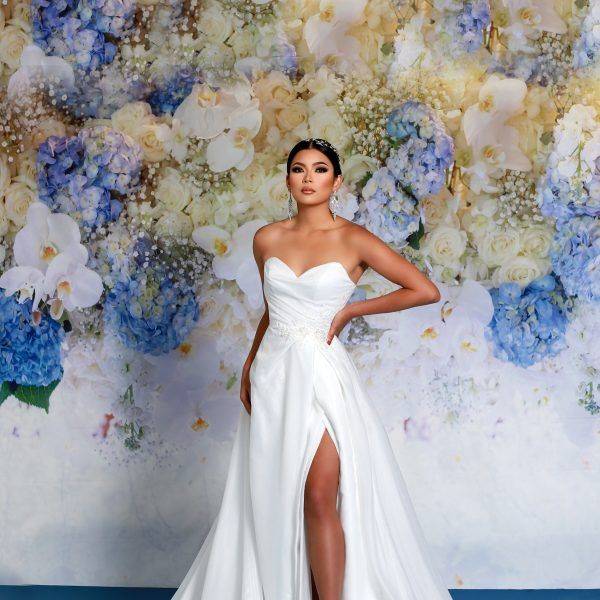 White satin gown with a deep side slit and sweetheart neckline, shown in front of a blue floral backdrop.