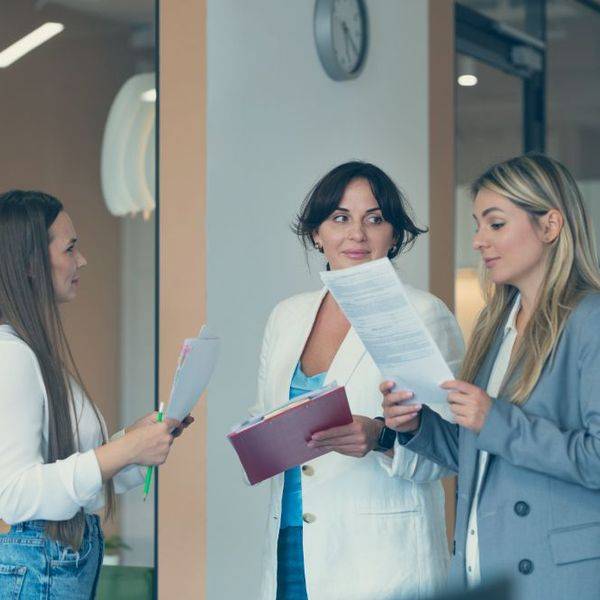 Three women reviewing paperwork together and talking in a bright, modern office.