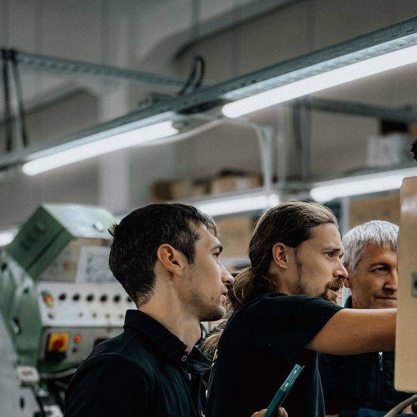 Two workers in black shirts looking at a handheld device beside industrial machinery.