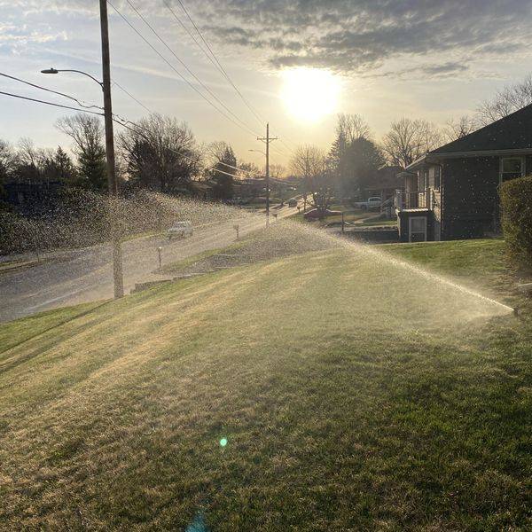 Lawn sprinkler spraying water across grass along a street at sunset.
