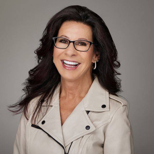 Smiling woman with dark hair and glasses in a beige jacket, against a light backdrop.