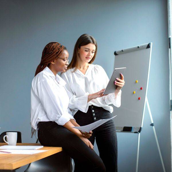 Two women review notes beside a flip chart in a bright office.
