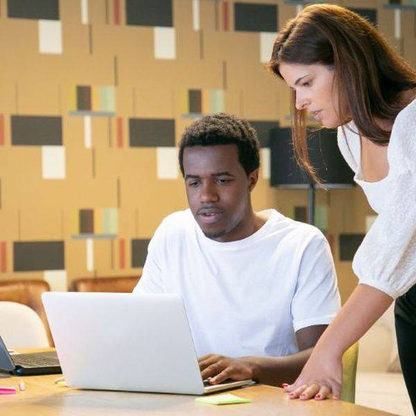 Two people work at laptops while a colleague leans in to help.